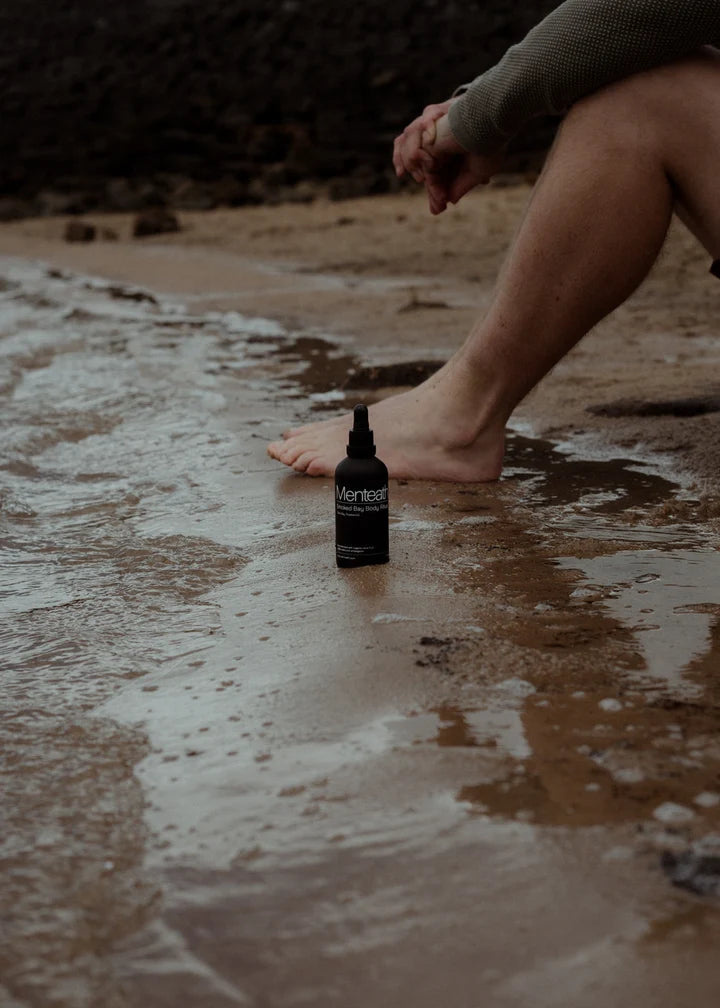 A Menteath bottle standing in wet sand at the water's edge beside a bare foot